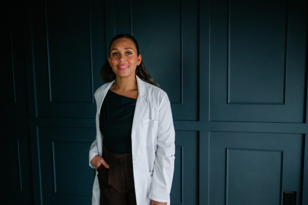 Portrait of a nurse injector in a white lab coat standing in front of a dark paneled wall