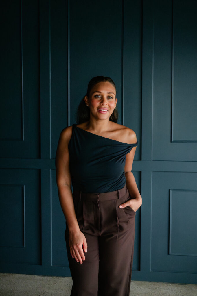 Standing portrait of a nurse injector in a one-shoulder top against a dark paneled wall