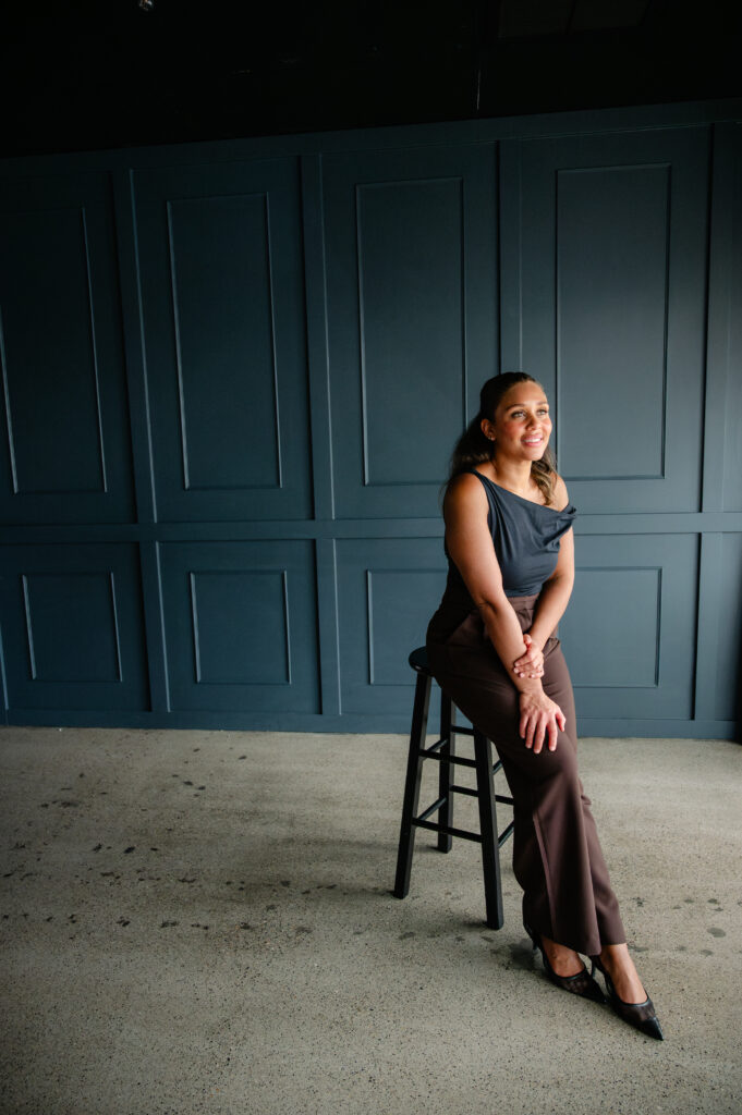 seated portrait of a woman in a one-shoulder top against a dark paneled wall