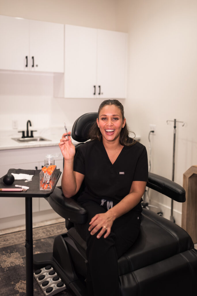 Aesthetic provider sitting in a treatment chair holding an injector syringe in a modern clinic room