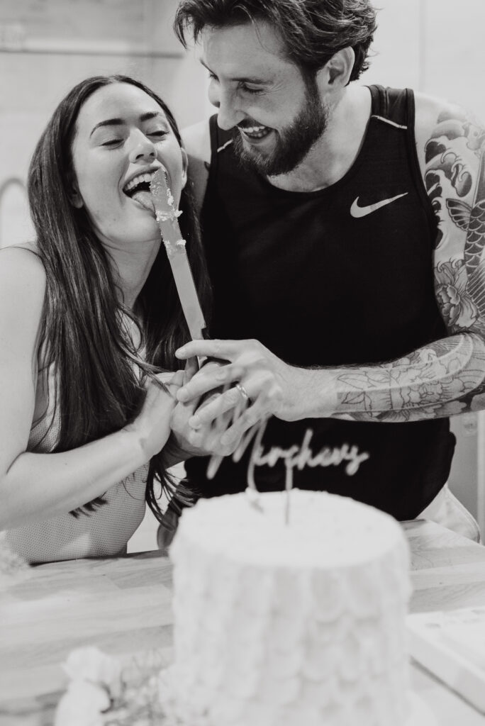 Black and white photo of couple laughing while cutting their elopement cake at home
