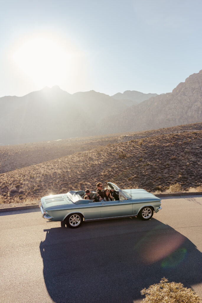Family riding in a blue vintage convertible on the Red Rock Scenic Loop at golden hour.