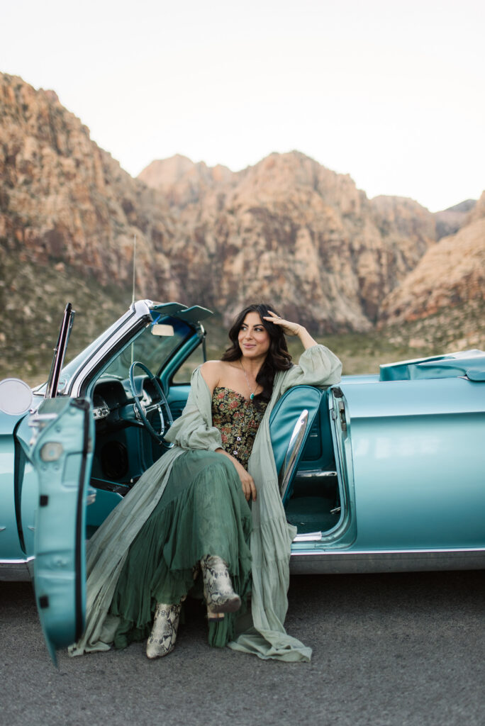 Woman posing in a blue vintage convertible with Red Rock Canyon cliffs behind her.