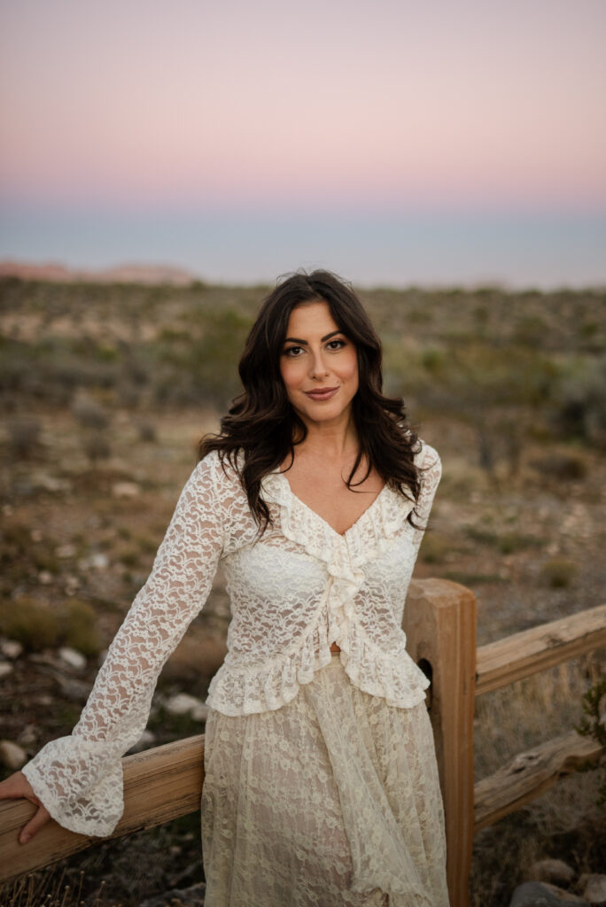 Woman in a white lace outfit leaning on a wooden railing at sunset in the desert.